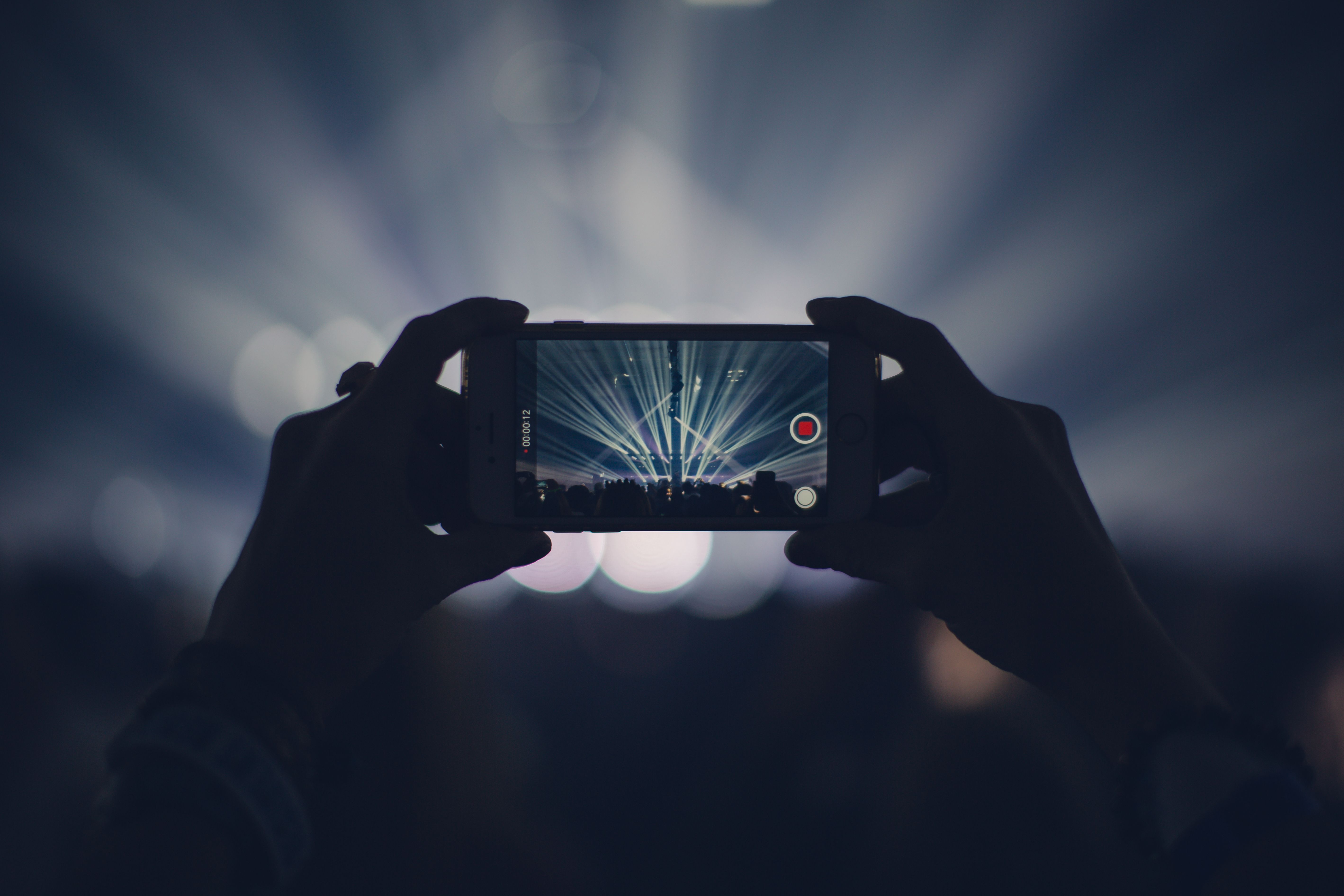 Hands holding a smartphone to record a brightly lit live performance, evoking real-time event coverage and audience engagement.