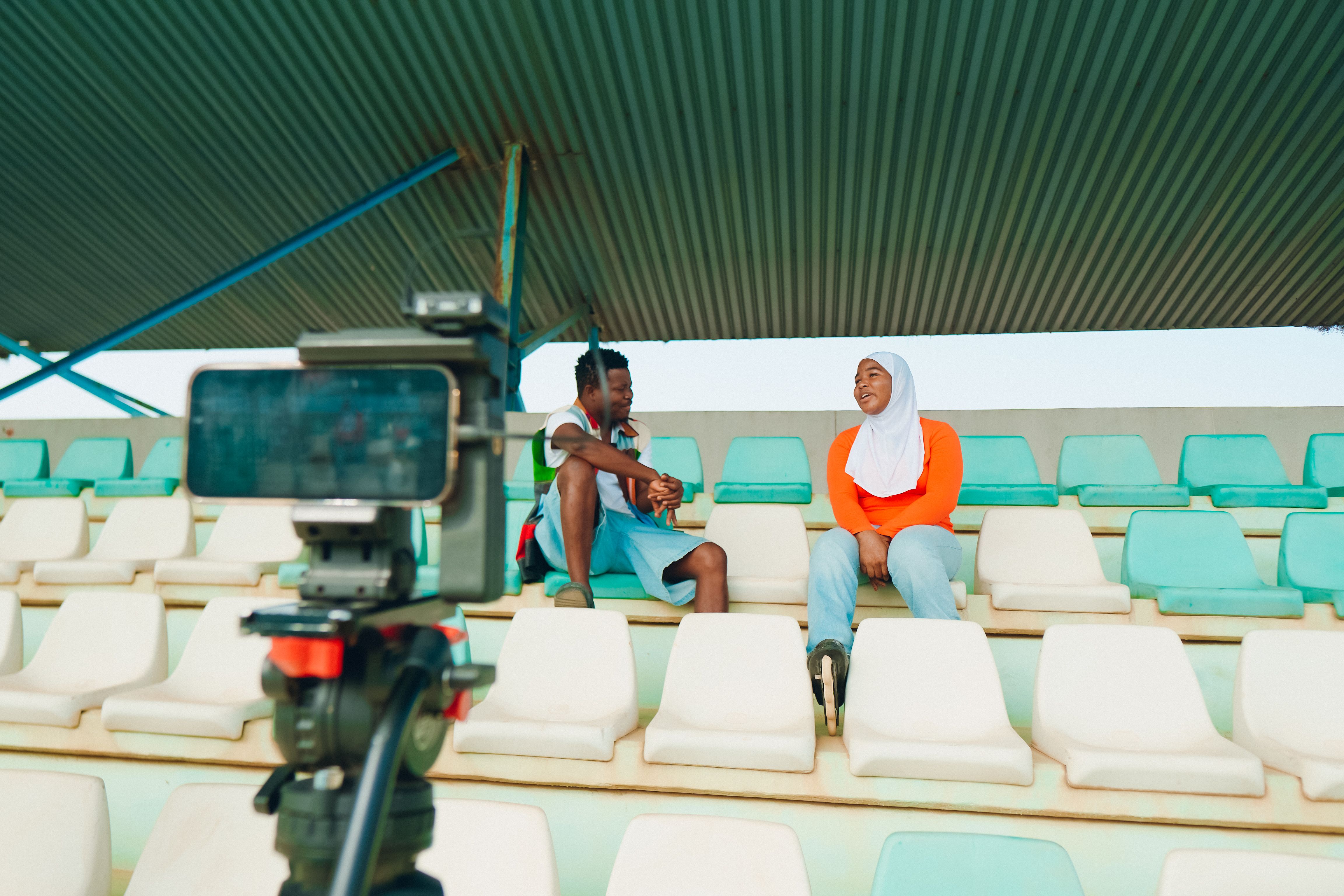 Two people conduct an interview in stadium seats with a video camera setup, illustrating behind-the-scenes sports content production.
